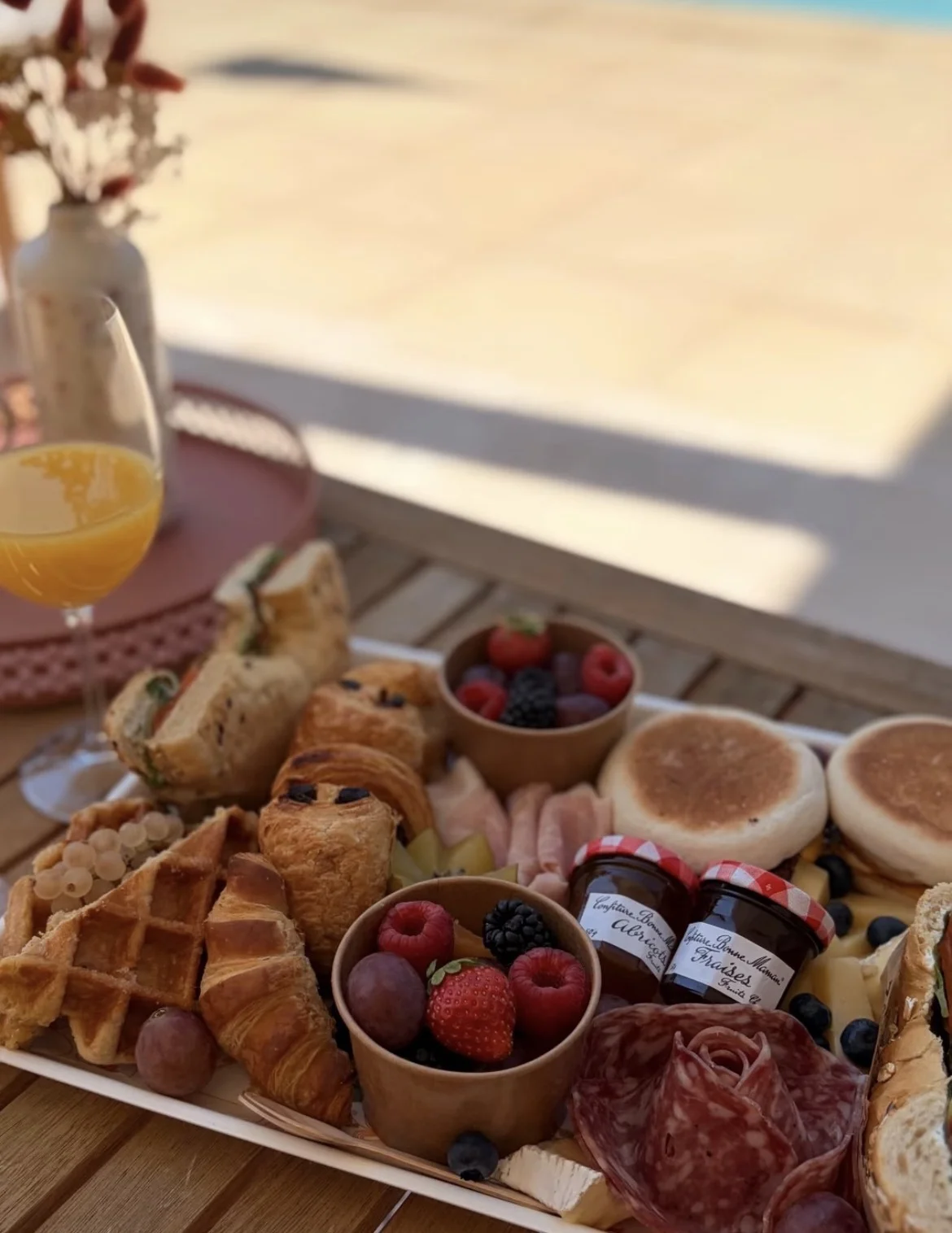 Plateau de la Brunch Box en extérieur : viennoiseries, sandwich, gaufre, fruits frais, charcuterie, fromages et confitures, avec un verre de mimosa